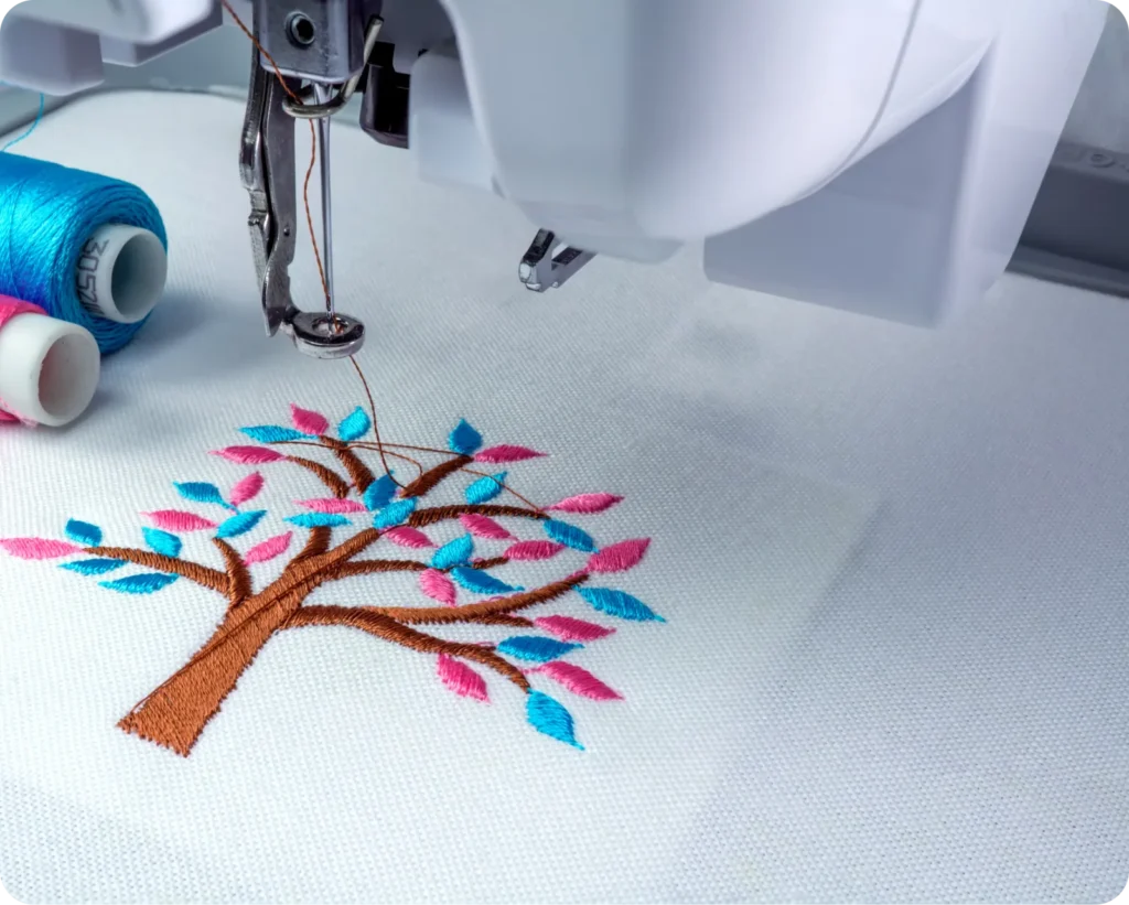 Sewing machine embroidering a tree with blue and pink leaves on fabric, with spools of blue, white, and pink thread nearby.