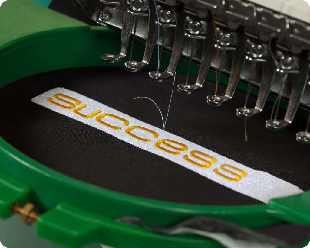 Machine embroidery in progress, stitching the word "success" in yellow thread on black fabric.